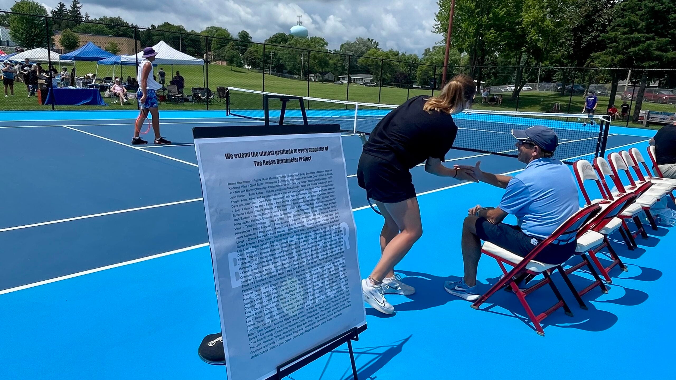 A sign on a stand is in the foreground near outdoor tennis courts, where players and a seated official interact. Several tents and people are visible in the background.
