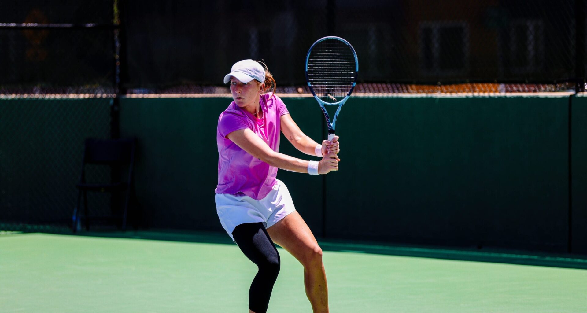 A tennis player in a pink shirt and white shorts prepares to hit a backhand shot on an outdoor court during daylight.