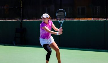 A tennis player in a pink shirt and white shorts prepares to hit a backhand shot on an outdoor court during daylight.