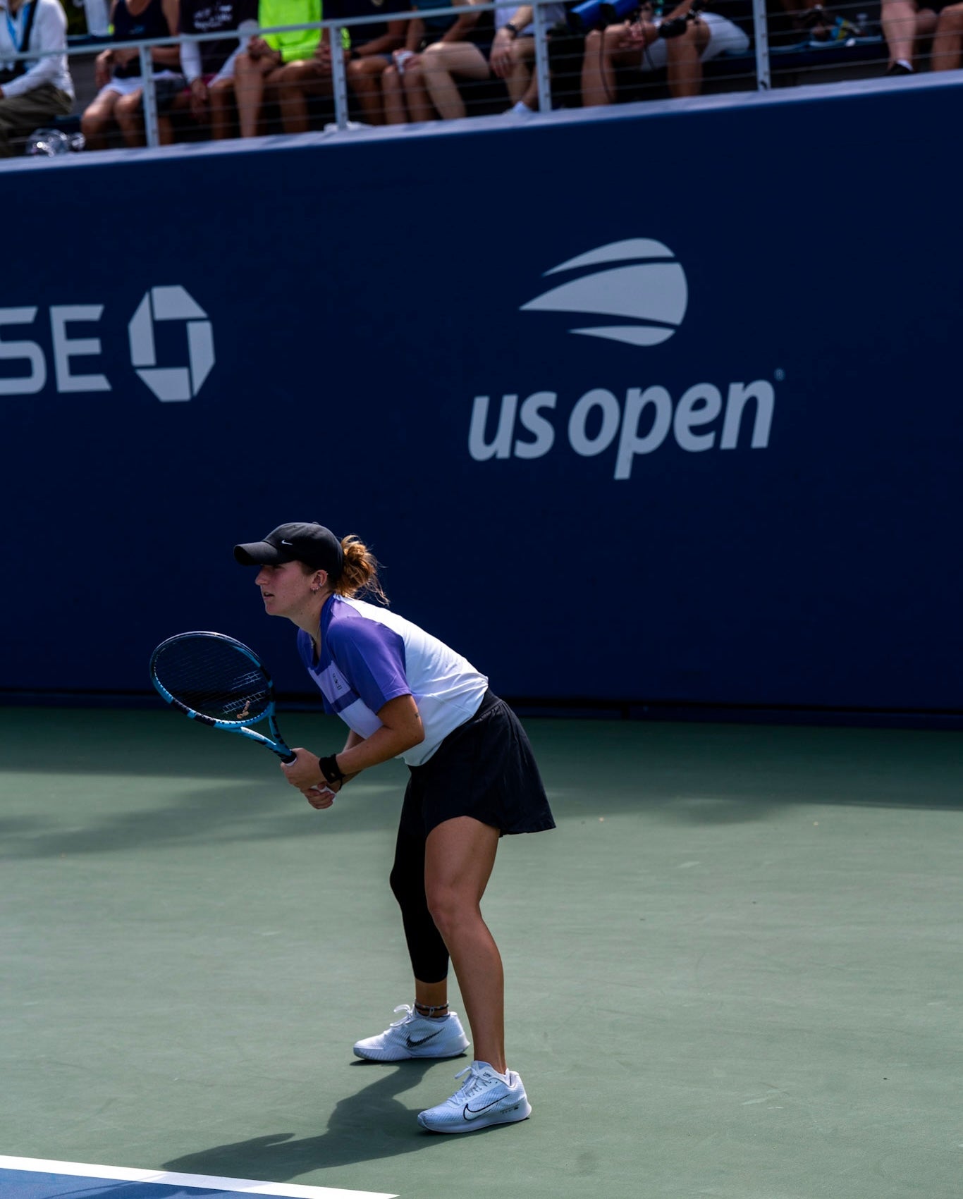 A tennis player stands ready to receive a serve on an outdoor court during a US Open match, with spectators in the background.