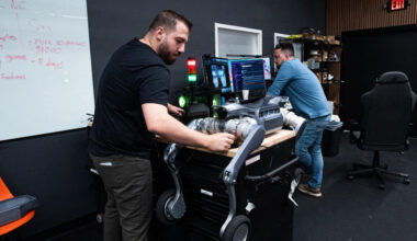 A person works on a quadruped robotic dog that is lying on a counter, while a second person stands in front of a computer with multiple display screens inside the office of Robo Inc.