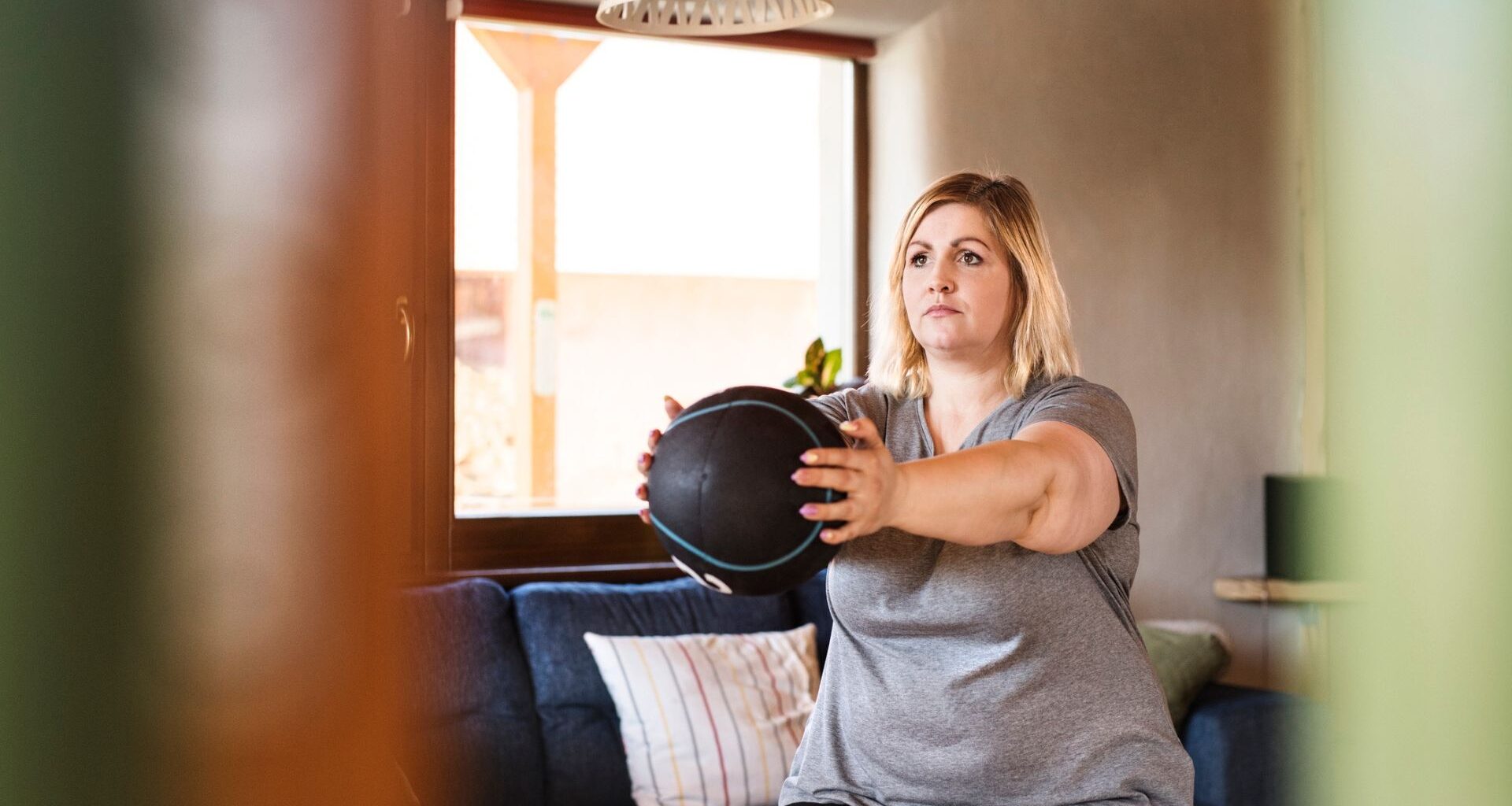 woman facing off camera standing holding a medicine ball arms outstretched in front. she's in a living room setting with a window behind her