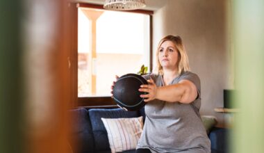 woman facing off camera standing holding a medicine ball arms outstretched in front. she's in a living room setting with a window behind her