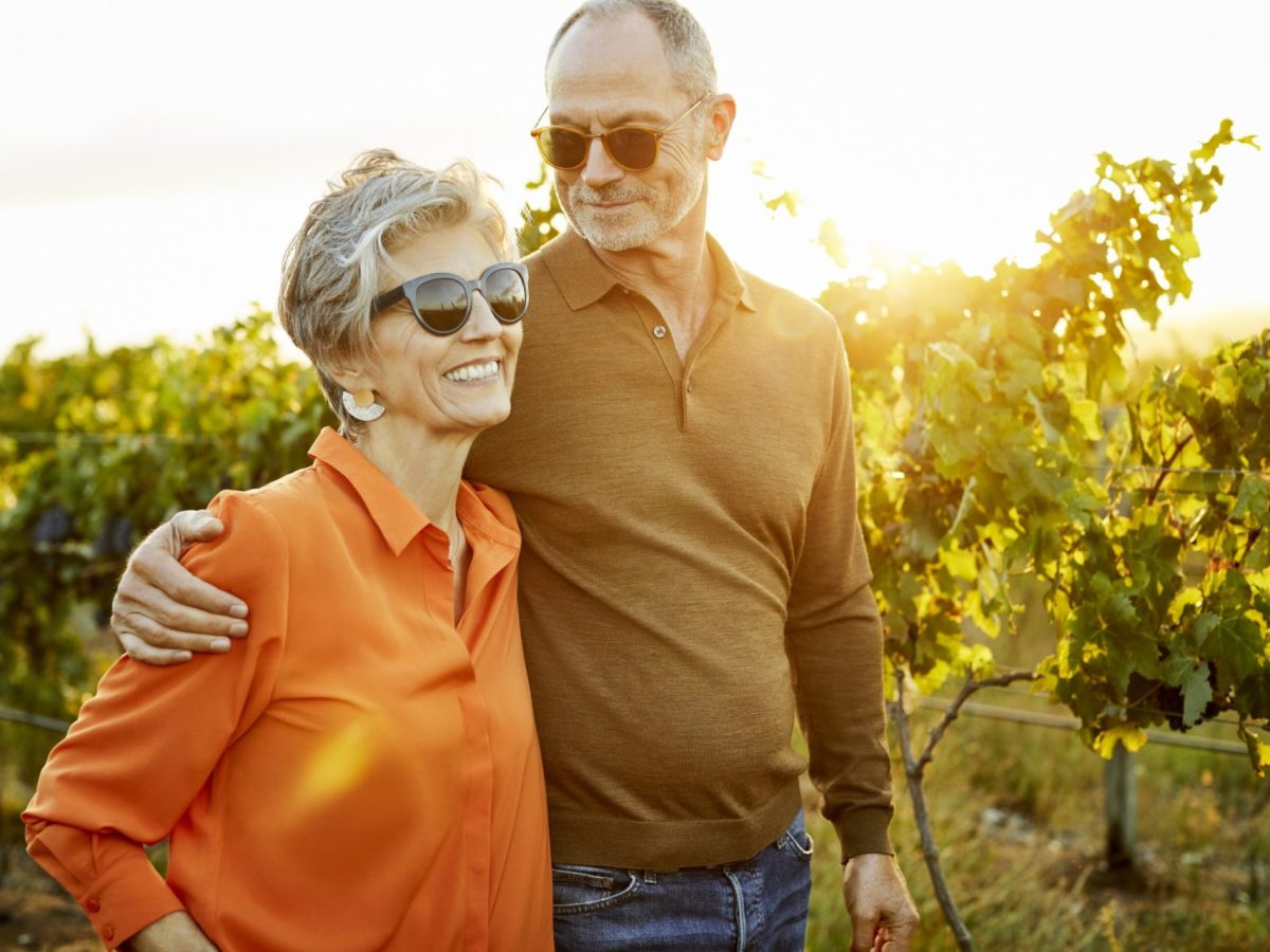 Happy woman walking with male partner in vineyard during sunset. Elderly couple are touring in field against sky. They are enjoying during weekend.