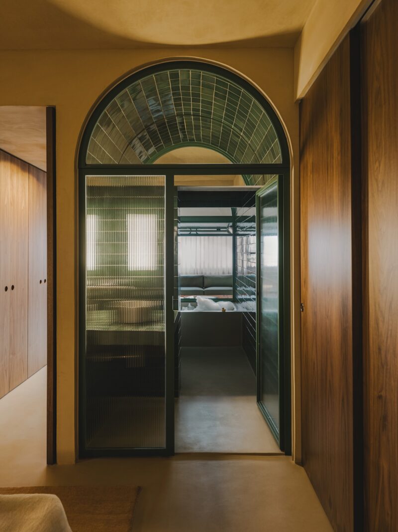 A glass door with a green frame and arched window opens to a room with bunk beds. Walls are wood-paneled; the floor is beige. The scene is softly lit with natural light.