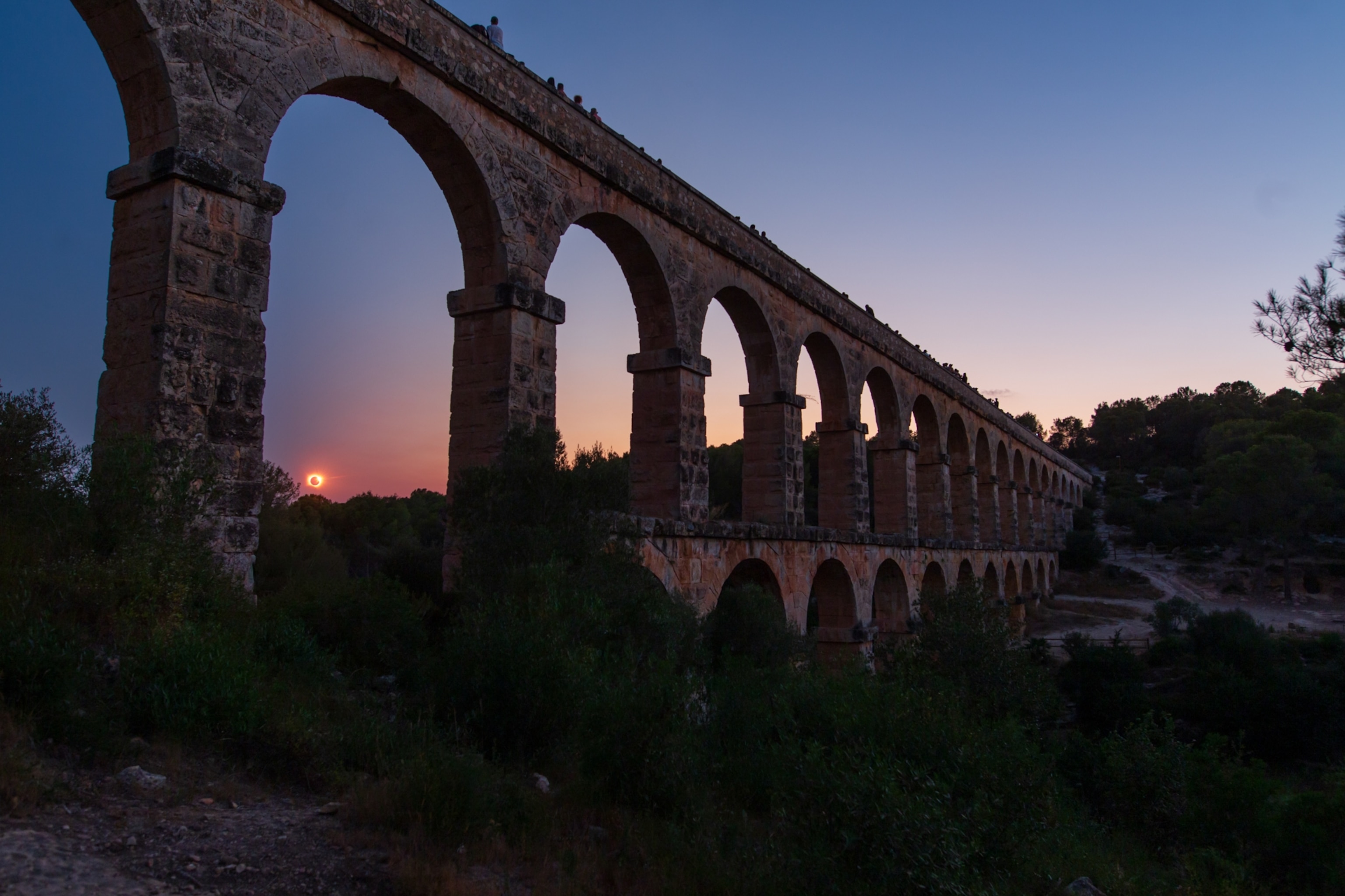 Ancient Roman stone aqueduct with repeating arches stretches through greenery during a solar eclipse, silhouetted against a pink and blue sky, serene.