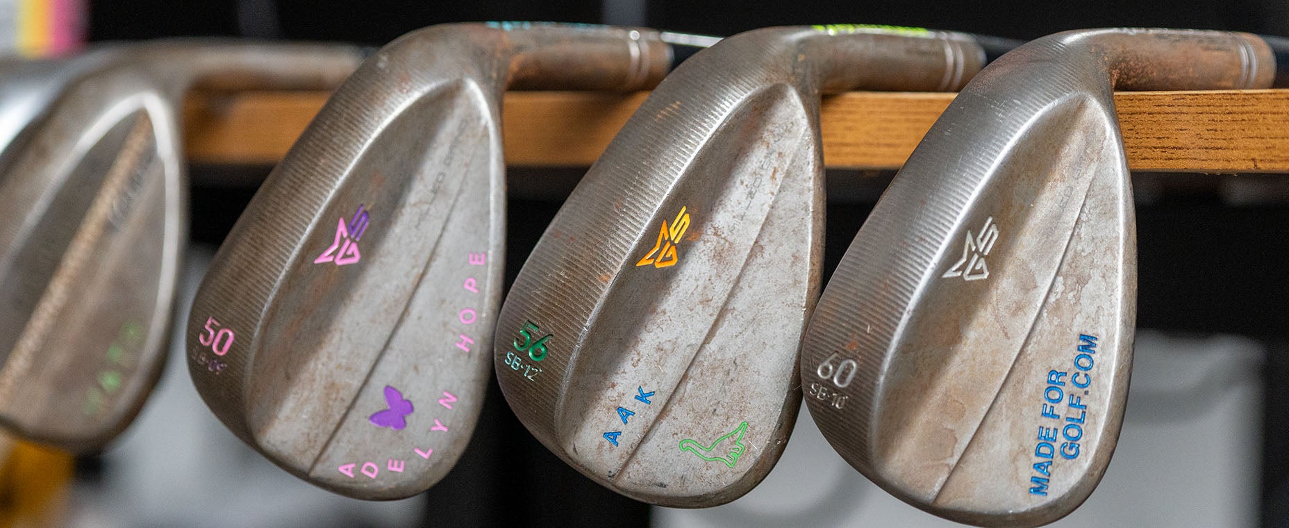 Three metal golf wedges with engraved, colorful text and initials rest side by side on a wooden rack. The clubs display numbers, symbols, and personalized engravings in pink, green, yellow, and blue—crafted to be your perfect wedges for a Fully Fit 2026 game.
