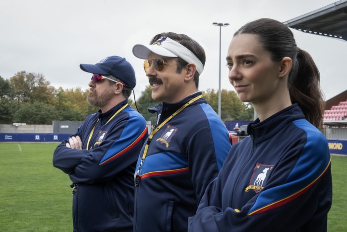 Ted Lasso Brendan Hunt, Jason Sudeikis and Tanya Reynolds star in Ted Lasso season 4; their characters are stood in a row on the edge of a football field, looking out at the players