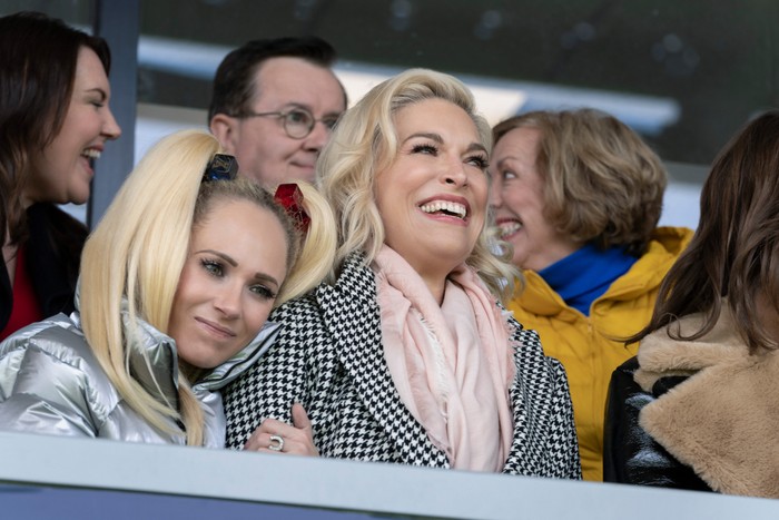 Ted_Lasso_Photo_031206 Keeley Jones, played by Juno Temple, rests her head on Rebecca Welton's shoulder as Rebecca, played by Hannah Waddingham, smiles while watching an AFC Richmond match from the stands. Other supporters sit around them in the stadium box.