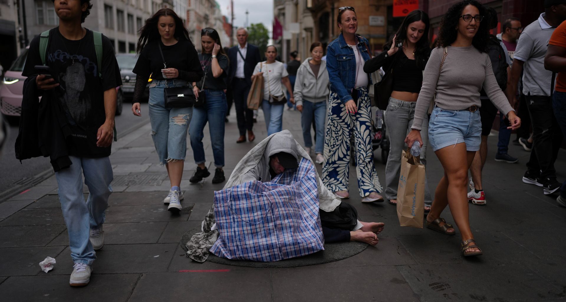 People rush past a man sitting on the street wrapped in a blanket.