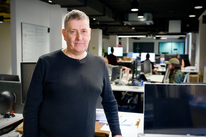 A man in a long black shirt stands in an open plan office with people and computers in the background.
