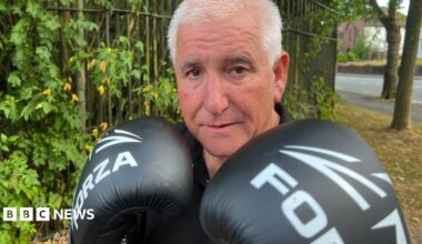 A man in a black T shirt is stood in front of a tree lined fence and looking directly into the camera. He is wearing boxing gloves which are raised to his chin in a fighting pose.