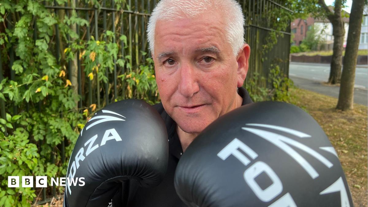 A man in a black T shirt is stood in front of a tree lined fence and looking directly into the camera. He is wearing boxing gloves which are raised to his chin in a fighting pose.