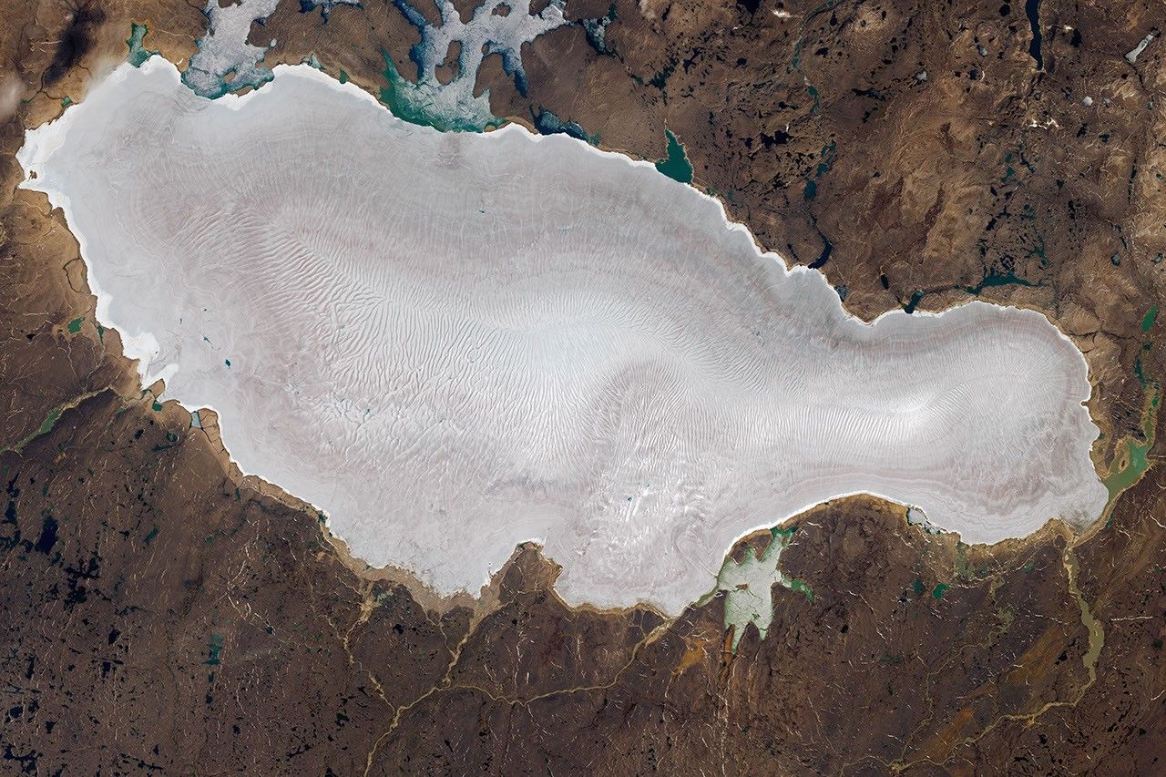 The surface of Barnes Ice Cap appears white, gray, and light brown. A fingerprint-like pattern of meltwater channels spans much of the ice. Brown land and several lakes, some ice-covered, surround it.