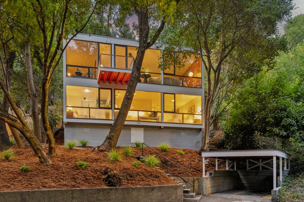 Modern two-story house with large windows, surrounded by trees and landscaping, featuring a patio and covered parking.