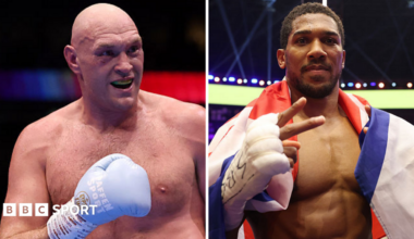 Tyson Fury holds up his hand and is wearing a boxing glove and Anthony Joshua holds up two fingers and has a Union Jack over his shoulders