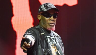 Dennis Rodman wearing sunglasses, a cap, and a leather jacket points forward on stage with a red background.