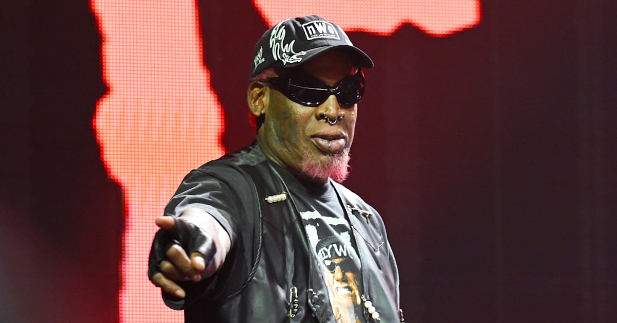 Dennis Rodman wearing sunglasses, a cap, and a leather jacket points forward on stage with a red background.