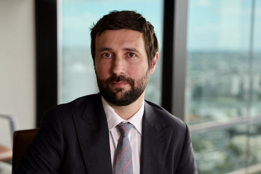 A man in a black suit and tie looks at camera.