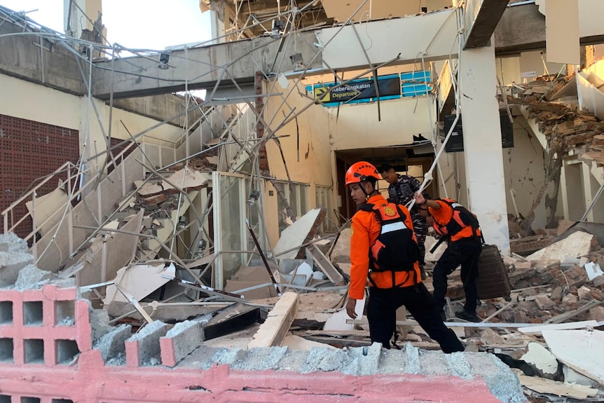 Two men in bright orange shirts and one man in military clothing look through rubble inside a building.