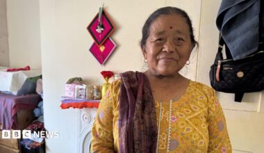 A Gurkha widow is wearing a bright yellow dress with a red shawl draped over her right shoulder. She also has a gold stud in her left nostril. Staring straight into the camera she's sitting in her bedroom and you can see the vague outlines of a few glass jars over her shoulder as well. A beautiful deep red shawl thrown over her right shoulder Indumati Thapa is standing in her small bedroom. Wearing a yellow dress you can see the foot stool she sits on when not in her bed behind her.
