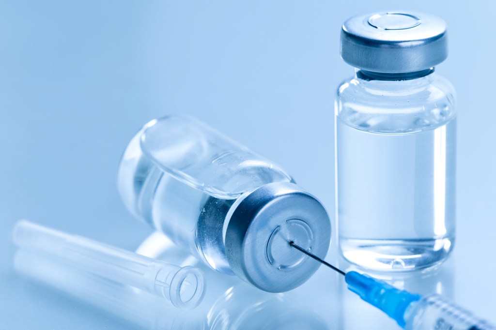 A medical syringe with a needle inserted into a vaccine vial, with another vial and a needle cap nearby.