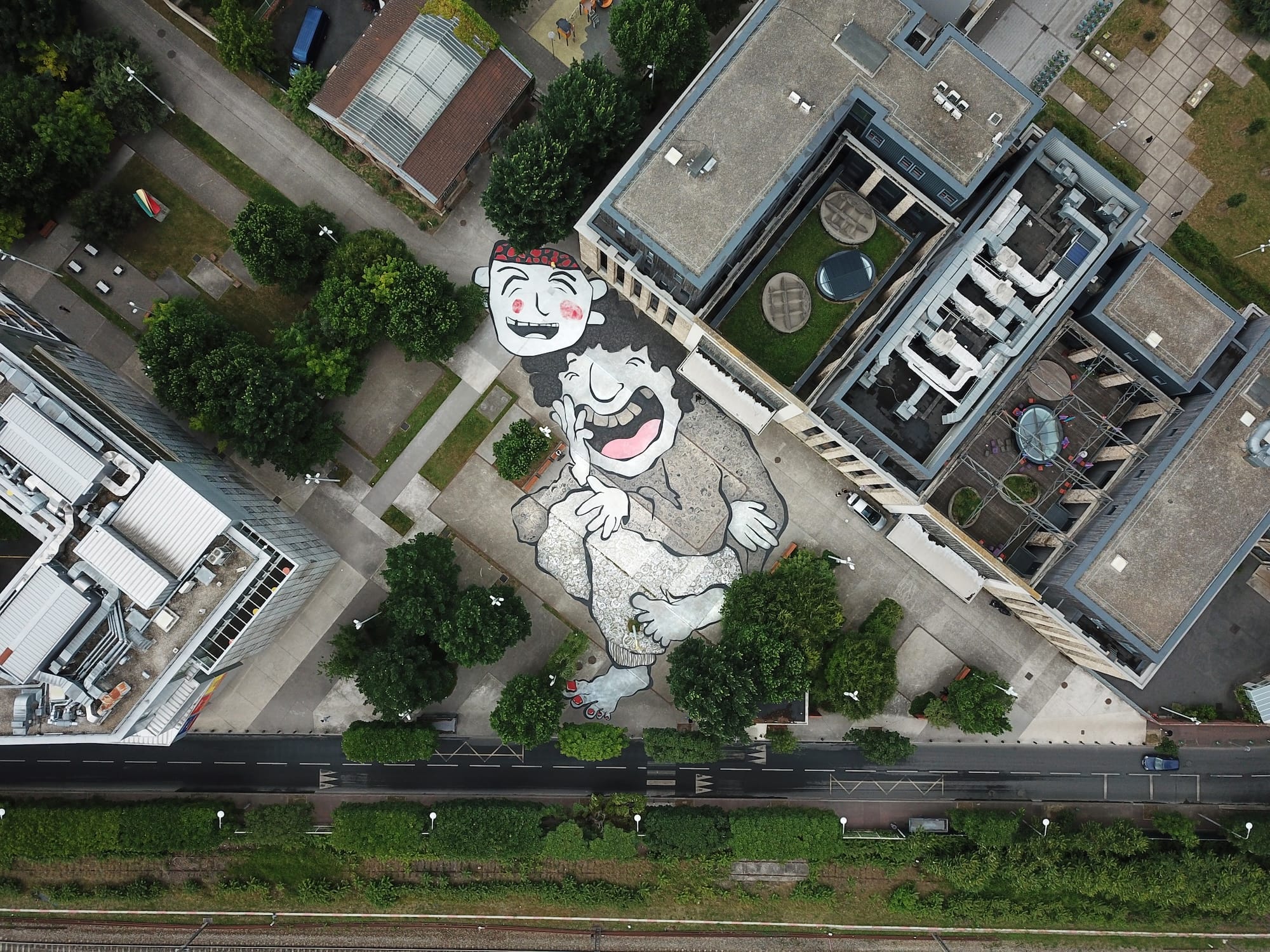 an aerial view of a large mural by Ella & Pitr on the ground of a public square featuring a cartoonish pair of laughing figures