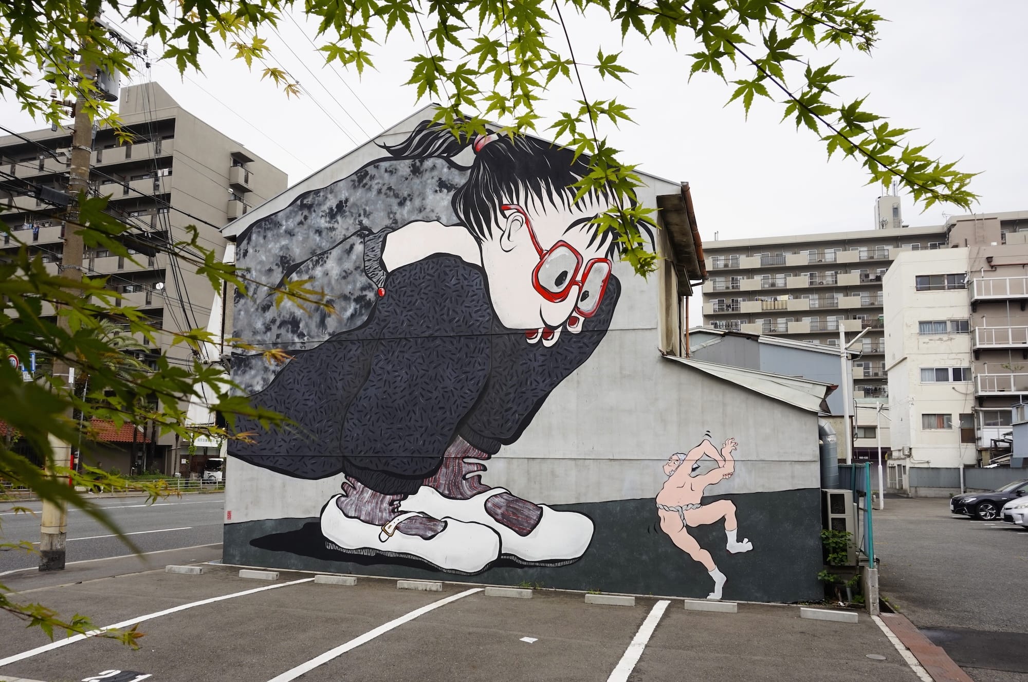 a mural by Ella & Pitr on a wall featuring two cartoonish figures, very large and looking down at a very small dancing figure without clothes on
