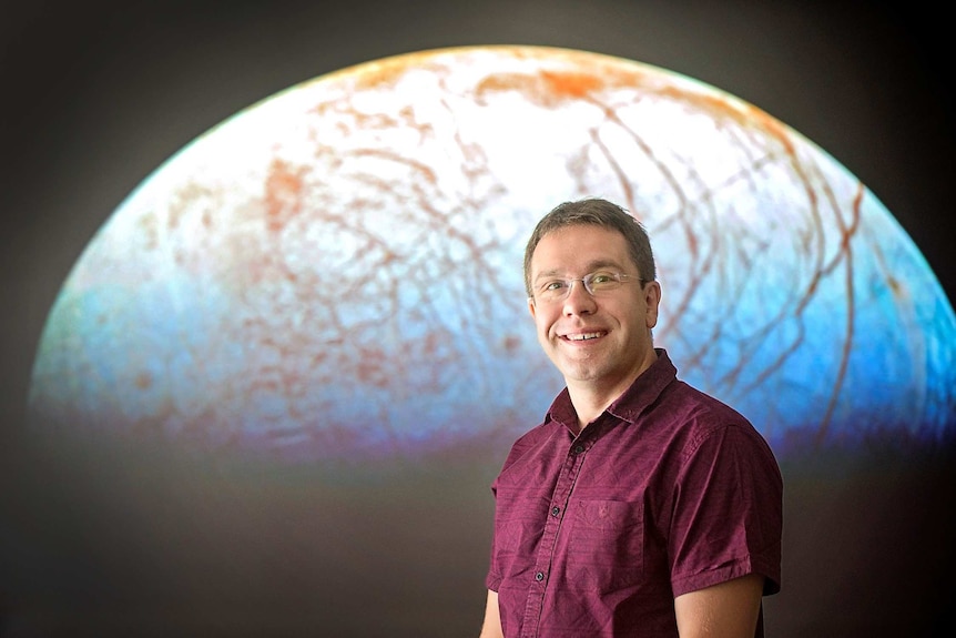 A man stands smiling in front of a background of the Earth.