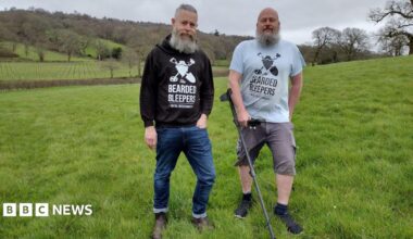 Two men with a metal detector in a field of green grass.