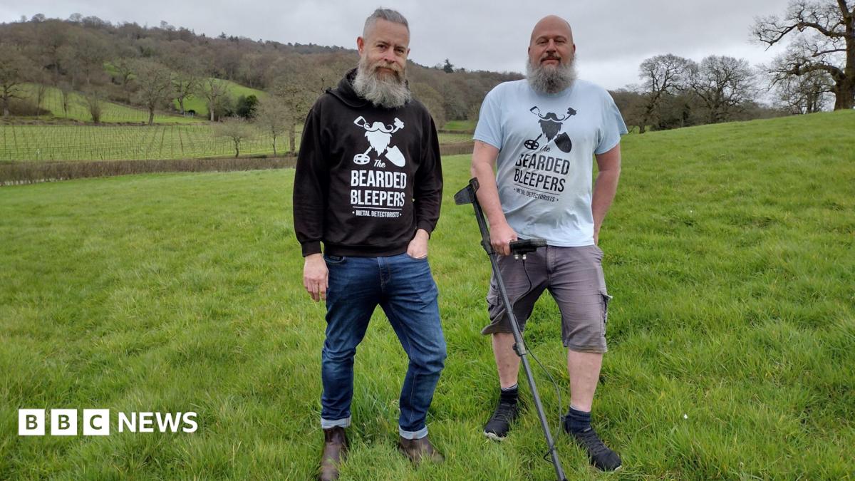 Two men with a metal detector in a field of green grass.