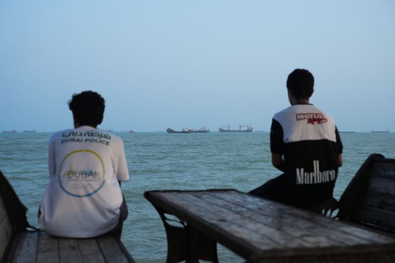 Two youths sit on the shoreline as ships are seen anchored in the Strait of Hormuz on August 10, 2026, off the coast of Bandar Abbas, Iran