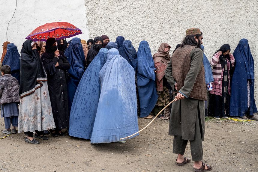 A member of Taliban security personnel stands alongside women who wait in a queue for food assistance in Kabul on March 25, 2025.