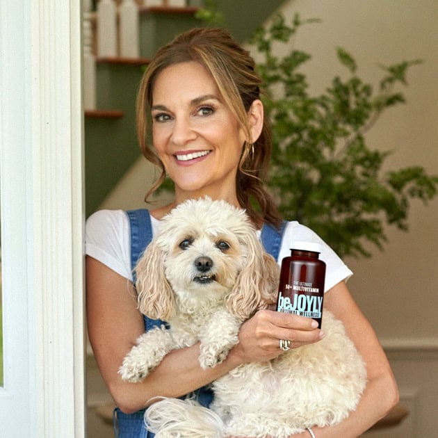A smiling woman stands in a doorway holding a small fluffy dog and a bottle labeled “BeJolly” supplement. A staircase and green plant are visible in the background.