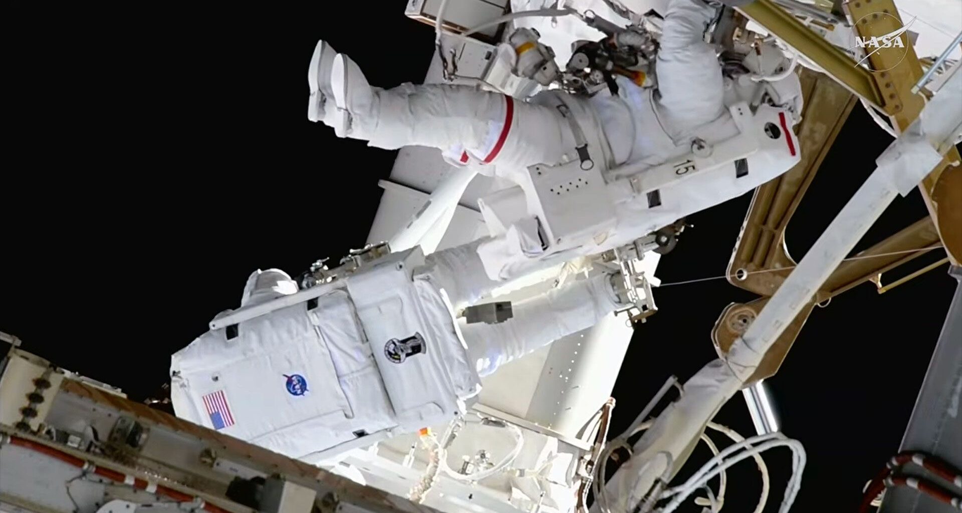Two astronauts in white bulky spacesuits work outside a large space station set against the blackness of space.