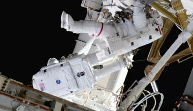 Two astronauts in white bulky spacesuits work outside a large space station set against the blackness of space.