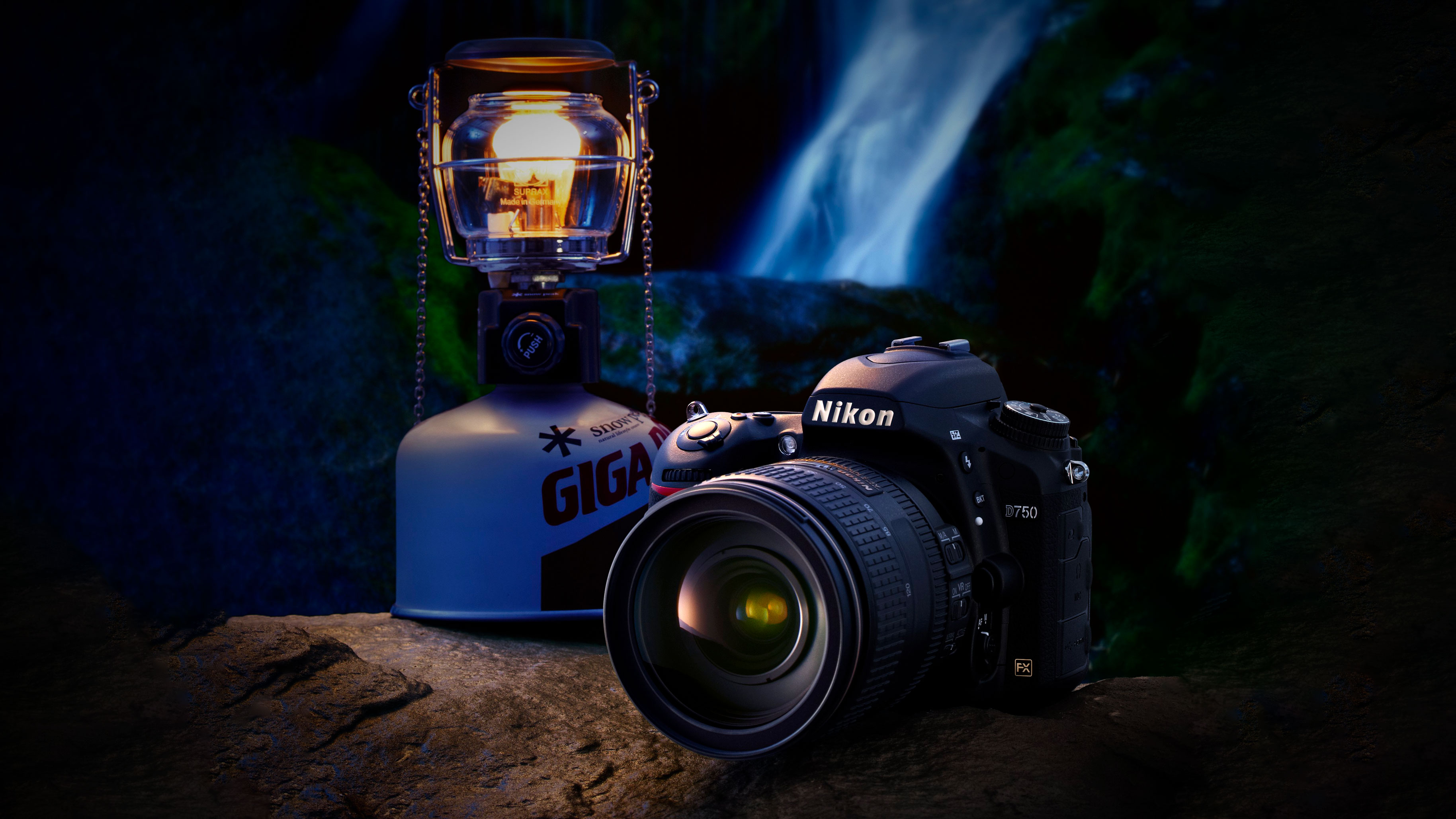 Nikon camera and lit camping lantern placed on a rock with a waterfall and dark forest background at night