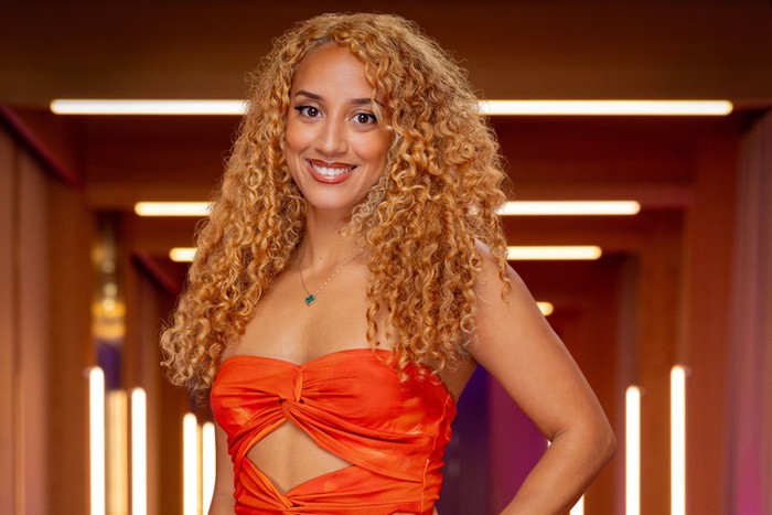 [A woman smiling ahead with one hand on her hip. She is wearing an orange bandeau dress and has curly ginger hair.]