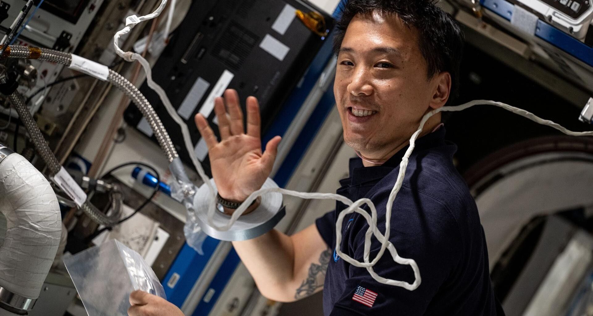 a man smiles and waves to a camera in a crowded laboratory full of wires and computer monitors