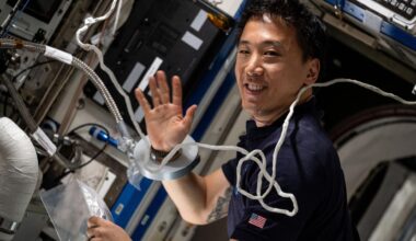 a man smiles and waves to a camera in a crowded laboratory full of wires and computer monitors