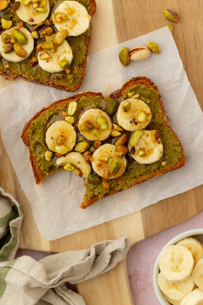 Two slices of toast topped with green pistachio spread, banana slices, chopped pistachios, and a drizzle of honey, arranged on parchment paper. One toast has a bite taken out. A bowl of banana slices is nearby.