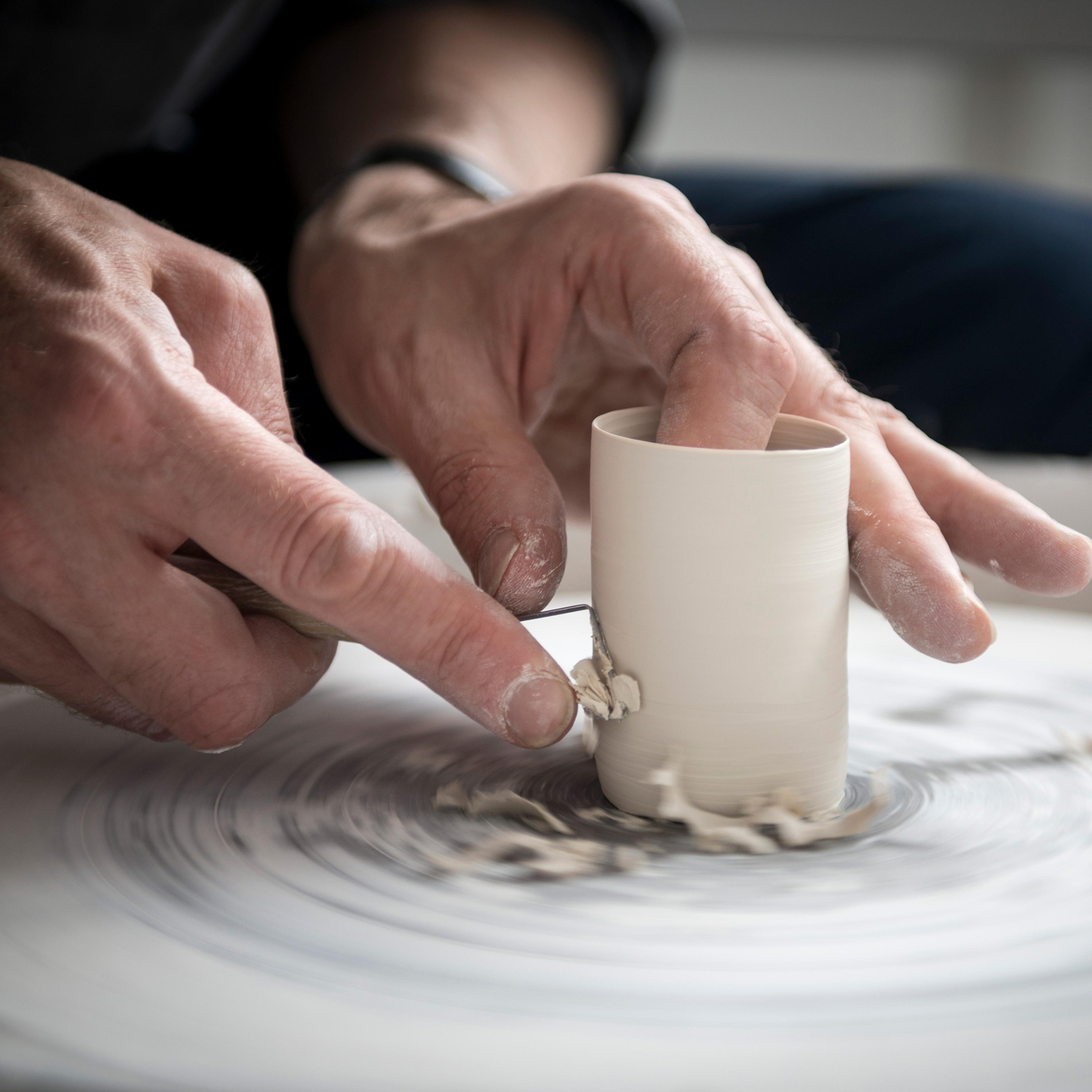 Hands shaping clay on a spinning pottery wheel, with a tool used for carving.