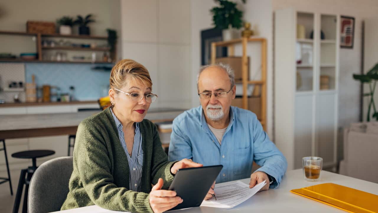 Senior couple sitting at a table and talking while looking at a tablet screen.