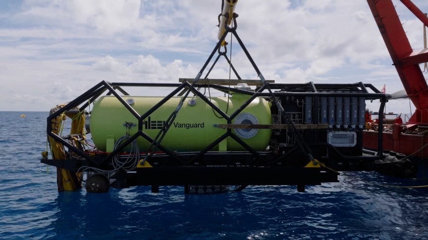DEEP's Vanguard habitat, a large yellow pressurized container, hangs from a crane over the ocean, about to be dropped into position on the sea floor.
