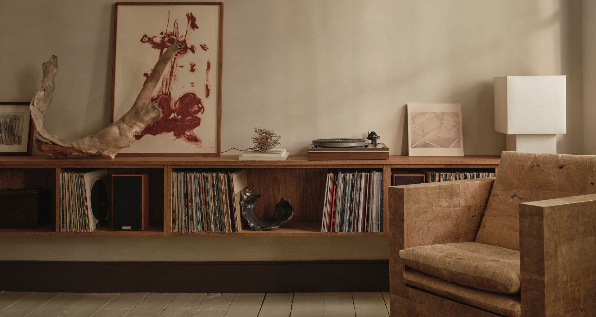A living room with a textured accent armchair, a floating display of vinyl, a record player, a white table lamp, a leaning painting, and ceramics
