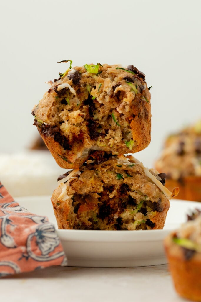 Two zucchini chocolate chip muffins are stacked on a white plate. The top muffin is broken open, showing its moist interior with green zucchini pieces and dark chocolate chips. A patterned orange napkin is on the left.
