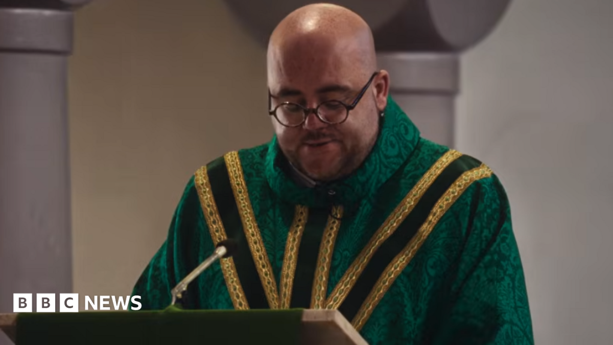 Father Luke wears a green chasuble with yellow brocade. He has a shaved head and dark round glasses.
