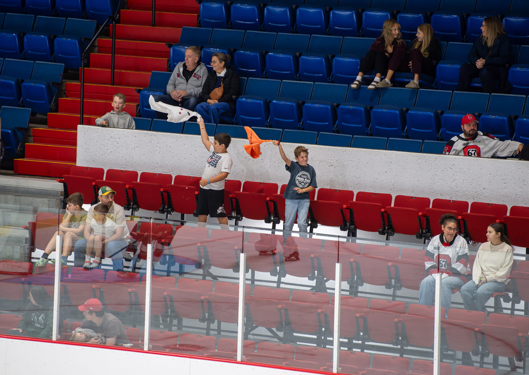 Two boys swinging shirts in the air in the stands at a hockey game.