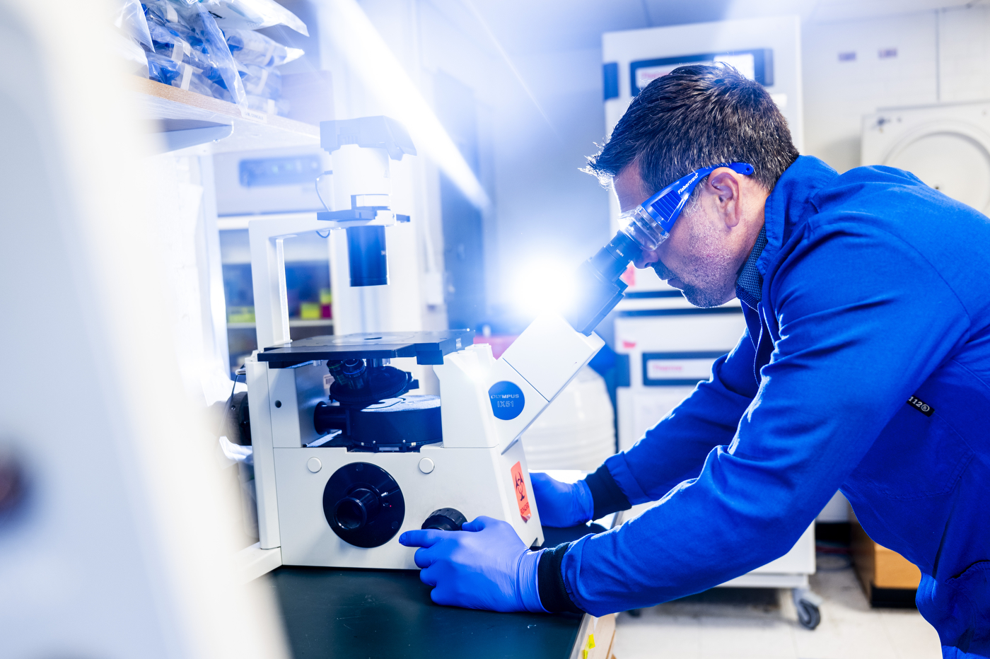 A researcher in a lab coat and goggles operates a laboratory instrument under blue light.