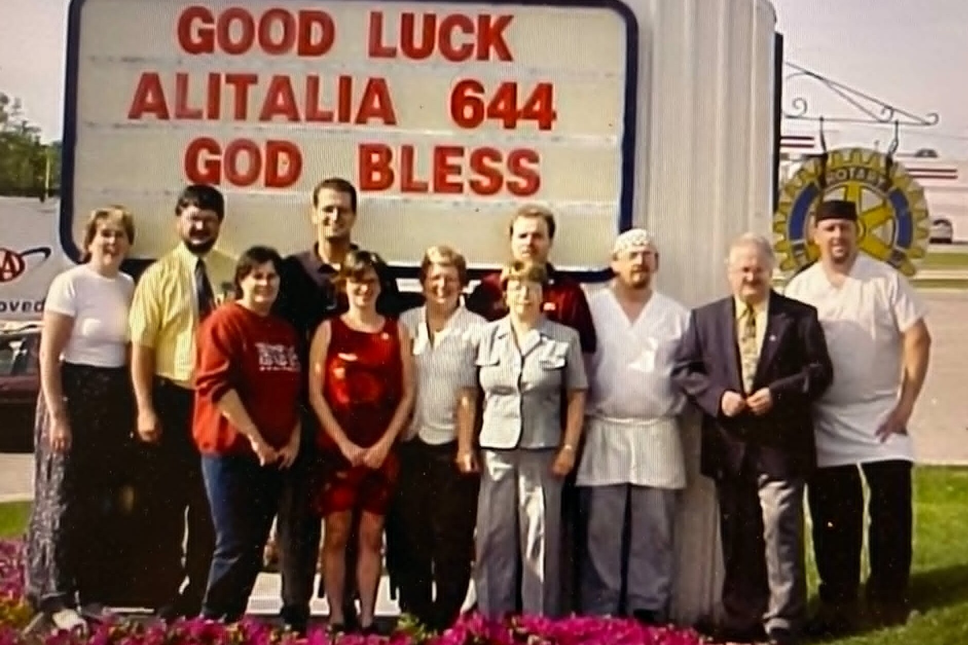 A group of people stand under a large sign that says GOOD LUCK ALITALIA 644 GOD BLESS